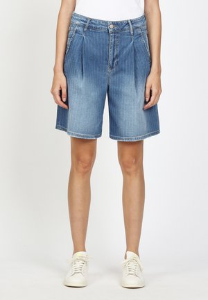 Person trägt kniehohe, blau gestreifte Jeansshorts und weiße Turnschuhe und steht vor einem schlichten weißen Hintergrund.