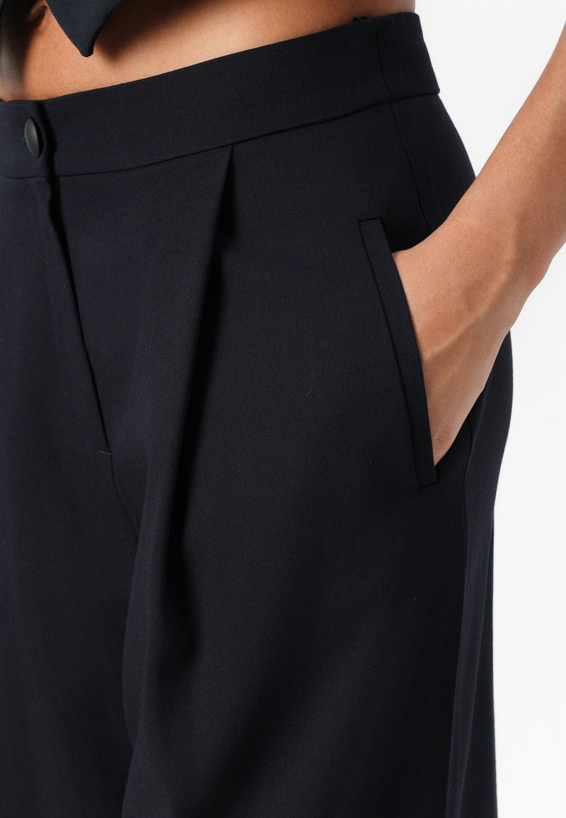 Maatwerk marineblauwe broek met een vlakke voorkant, zijzakken en een gladde, gestructureerde stof. Voorzien van een knoopsluiting en lichte plooien.