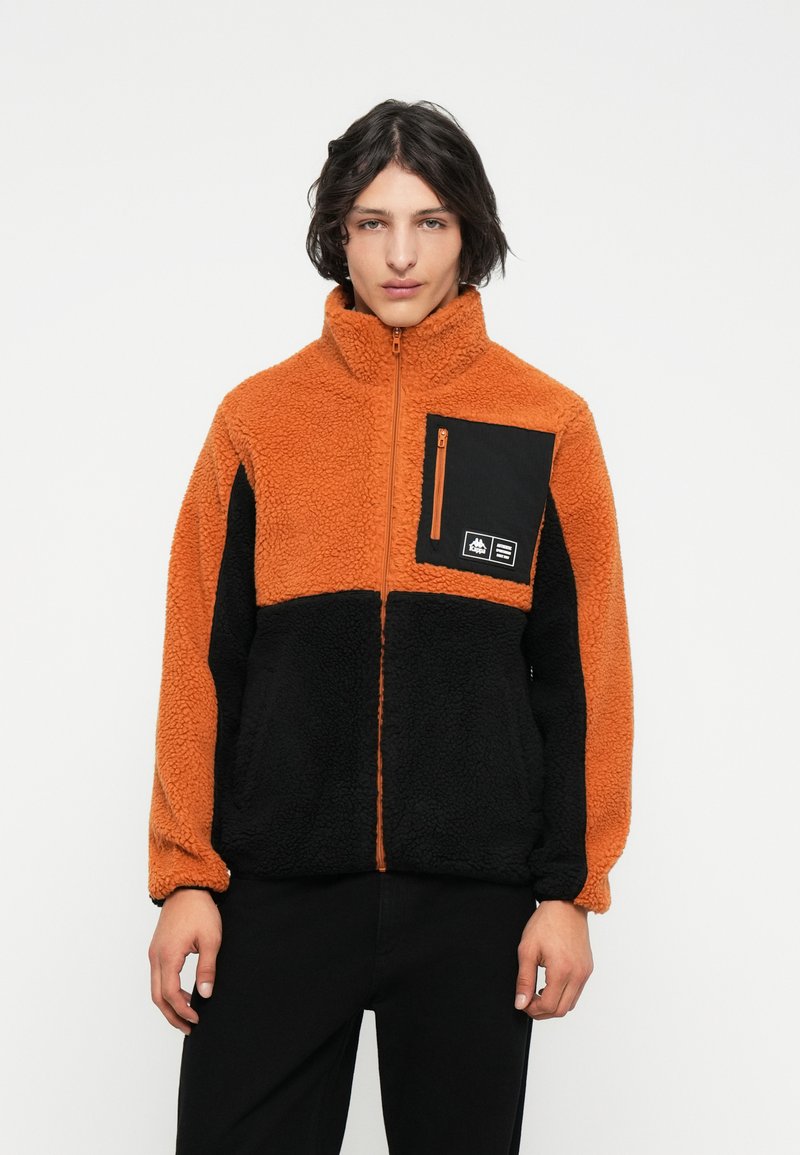 Chaqueta de forro polar con secciones en naranja y negro, cuello alto, cremallera frontal y un bolsillo en el pecho con detalles de metal y marca.