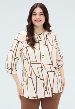 Femme souriante aux longs cheveux foncés portant un chemisier crème avec des lignes abstraites marron et un pantalon marron, debout devant un fond clair.