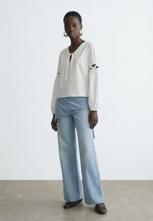 Donna che indossa una blusa bianca a maniche lunghe con dettaglio a fiocco e jeans azzurri a gamba larga con sovrapposizione di denim asimmetrico, in piedi su pavimento grigio.