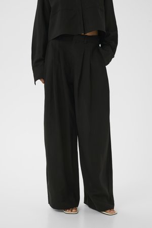 Person trägt hoch taillierte, weit geschnittene schwarze Hose mit einem schwarzen, kurzen Hemd mit Knöpfen und beigefarbenen Sandalen mit quadratischer Zehenpartie.