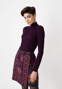 Lila gerippter Rollkragenpullover kombiniert mit einem gemusterten Minirock, der mehrfarbige Blumen- und geometrische Designs auf einem dunklen Hintergrund aufweist.