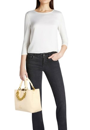 Borsa beige con finitura testurizzata, dettagli in catena dorata e due manici, abbinata a una maglia bianca a maniche lunghe e jeans neri.