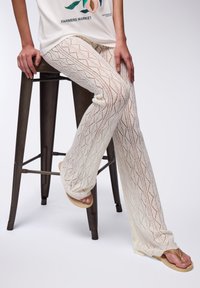 Pantaloni in pizzo crema a maglia con un motivo ondulato, abbinati a infradito piatte color cuoio, modello seduto su uno sgabello di metallo su uno sfondo chiaro.