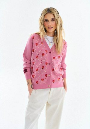 Kobieta o długich blond włosach nosi różowy sweter w wzory zapałek i płomieni, białą koszulę oraz kremowe spodnie, stoi z rękami w kieszeniach.