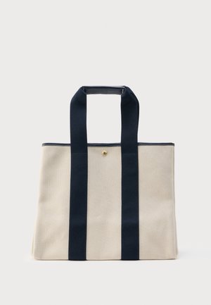 Beige canvas boodschappentas met twee brede zwarte banden die van de basis naar boven lopen en handvaten vormen, en een kleine gouden drukknopsluiting aan de bovenkant in het midden.