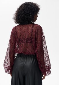 Przezroczysta bordowa bluzka z koronkowym wzorem i kropkami, z długimi, bufiastymi rękawami i czarnym kołnierzem, zestawiona z czarnymi skórzanymi spodniami.