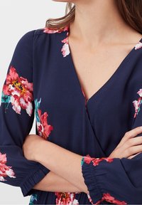 Květinové zavinovací šaty v námořnické modři s růžovými a zelenými květinami. Mají dlouhé rukávy s nabíranými manžetami a V výstřihem. Hladká textura tkaniny.