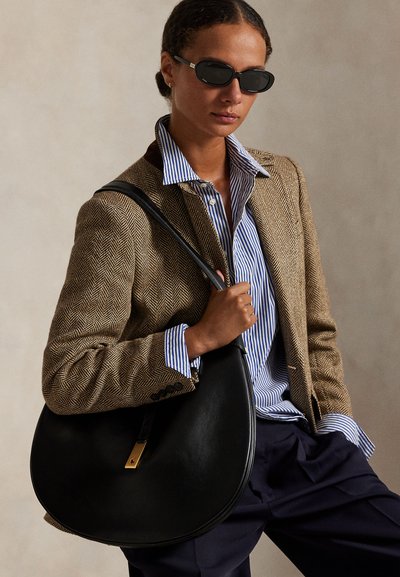 Femme portant un blazer marron en chevrons, une chemise bleue rayée, un pantalon foncé, des lunettes de soleil noires, tenant un grand sac à main en cuir noir sur l’épaule.