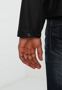 Svart vattentät jacka med knappstängning på ärmen, som visar en hand vilande vid manschetten. Jeansen är synliga i nedre högra hörnet.