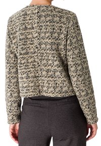 Personne portant une veste texturée beige et noire à motifs et un pantalon gris foncé, vue de dos, debout les mains relâchées le long du corps.