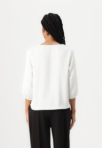 Žena se zkroucenými copy, oblečená v volné bílé blůze s třičtvrtečními rukávy a černými širokými kalhotami, viděná ze zad.