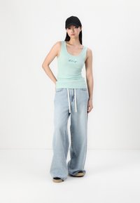 Débardeur à côtes vert clair avec "GAP" en lettres bleues, associé à un jean en denim bleu clair à jambes larges et une casquette noire. Tissu texturé et coupe ample.