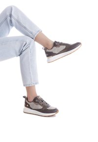 Zapatillas deportivas marrones y grises con paneles de malla, suela blanca gruesa y acentos de metal. Llevadas con jeans de color azul claro, mostrando un look casual.