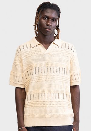 Mand med flettet hår iført en beige tekstureret strikket kortærmet polo, stående foran en ensfarvet baggrund.