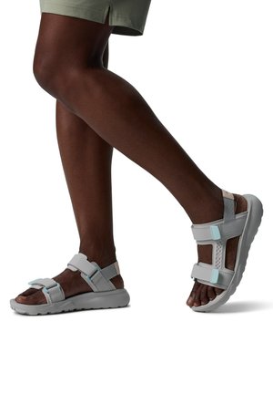 Dunkelhäutige Person trägt hellgraue sportliche Sandalen mit dicken Sohlen und blauen Akzenten, posiert mit einem leicht angehobenen Fuß.