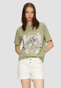 oliwkowa bawełniana koszulka z nadrukiem, luźny krój. W połączeniu z białymi szortami denimowymi z zapięciem na guziki i podwiniętymi nogawkami.