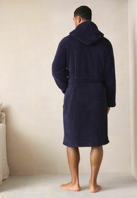 Granatowy szlafrok z kapturem, o teksturowanym wzorze, do kolan, materiał wydaje się pluszowy, z związanym w pasie i długimi rękawami.