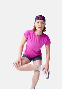 Ružová športová košeľa s krátkymi rukávmi, šedé šortky a biele tenisky. Osoba sa natiahne s jednou nohou, pričom má na sebe námornícku čiapku.