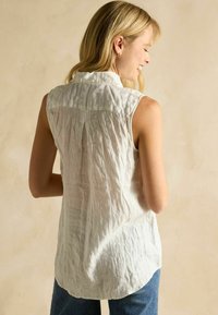 Cămașă fără mâneci din in alb, cu o textură ușor sifonată, guler și tiv curbat, purtată cu blugi din denim albastru.