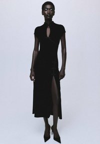Vestido negro ajustado con un cuello alto, mangas cortas y una abertura lateral. Presenta un detalle tipo ojal y acentos de botones. Combínalo con tacones.
