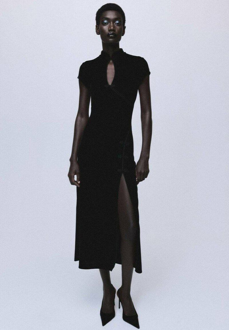 Vestido negro ajustado con un cuello alto, mangas cortas y una abertura lateral. Presenta un detalle tipo ojal y acentos de botones. Combínalo con tacones.