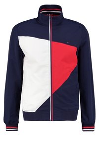 Marinblå jacka med ett vitt och rött geometriskt mönster, dragkedja, hög krage och ribbade ärmmuddar med randiga detaljer.