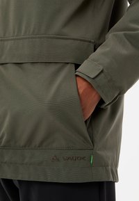 Giacca verde oliva con una texture liscia, dotata di una tasca laterale, polsini regolabili e un discreto marchio VAUDE lungo l'orlo.