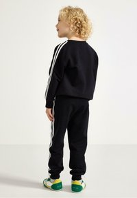 Černá sada mikiny a kalhot s bílými proužky na bocích. Vyrobena z jemné látky, má žebrované manžety a sportovní zelené, žluté a bílé tenisky.