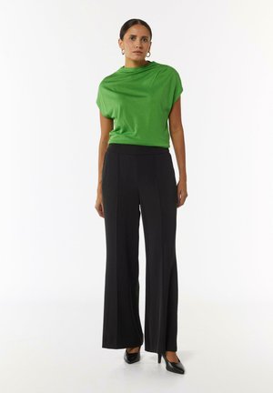 Grünes Kurzarmoberteil kombiniert mit hochgeschnittenen schwarzen weiten Hosen. Glatter Stoff, minimalistisches Design und schmal zulaufende Beine. Schwarze Absätze.