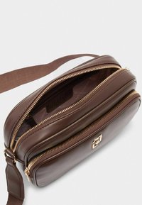 Hnědá kožená crossbody taška s dvojitým zipem, hladkou texturou, zlatým kováním a látkovým popruhem. Uvnitř se nachází logo značky.