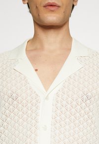 Cárdigan blanco de punto con un patrón de diamante y un cuello con solapa. Presenta una textura transparente y cuatro botones visibles.