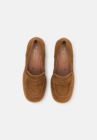 Et par brune ruskinds loafers med teksturerede gummisulder, syet detaljer og enrem over det øverste, vist ovenfra på en hvid baggrund.