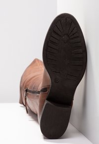 Botte en cuir marron posée sur le côté, montrant la semelle noire texturée et la fermeture éclair latérale sur fond blanc.