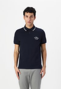 Marinepiké med krage, hvit kant og brodert logo. Har kort erm og vanlig passform. Et par lysegrå bukser.