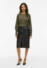 Vrouw in een olijfgroene blouse met lange mouwen, zwarte leren rok tot de knie met een split aan de voorkant, en zwarte hoge hakken met enkelbandjes.