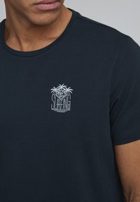Marineblå kortærmet t-shirt lavet af bomuld med et hvidt broderet surfinglogo med palmer og bølger. Rund halsudskæring.