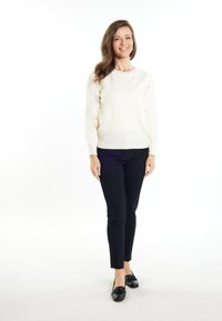 Pulover de culoare crem cu guler rotund și accente texturate, asortat cu pantaloni negri strâmți și mocasini negri. Modelul stă în fața unui fundal alb.
