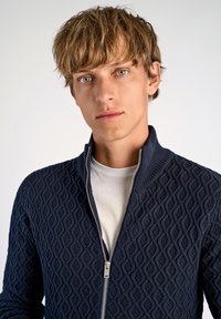 Marinblå zip-up kofta med en texturerad diamantmönster, med hög krage och silverdragkedja. Bärs över en vit t-shirt.