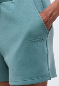 Το χέρι ενός ατόμου στην τσέπη μιας τιρκουάζ βερμούδας με διακριτικό ανάγλυφο λογότυπο "BORG" στην μπροστινή περιοχή του μηρού.