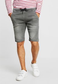 Pantalones cortos de mezclilla grises con cintura ajustable, con un acabado ligeramente desgastado. Llevados con una camisa de manga larga a rayas y zapatillas blancas.