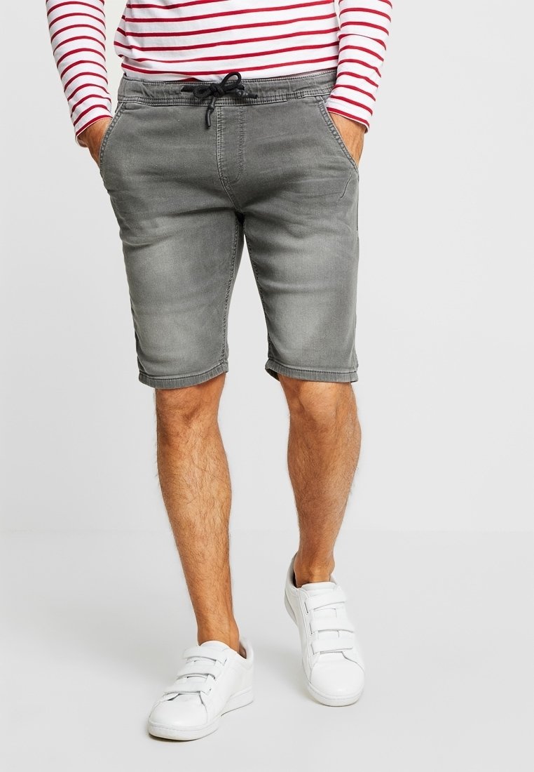 Pantalones cortos de mezclilla grises con cintura ajustable, con un acabado ligeramente desgastado. Llevados con una camisa de manga larga a rayas y zapatillas blancas.