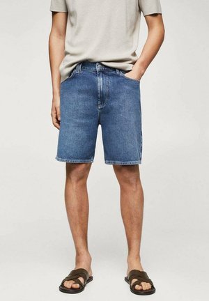 Mand iført blå denimshorts, en beige kortærmet skjorte og brune sandaler står med den ene hånd i lommen.