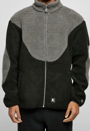 Hombre vestido con chaqueta de forro polar bicolor en negro y gris, con cremallera, cuello alto y tejido texturizado, posando frente a un fondo liso.