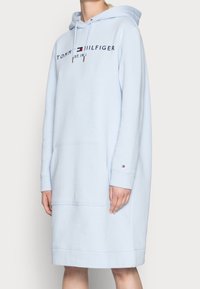 Lichtblauwe Tommy Hilfiger hoodie-jurk met een voorzak, logo-opdruk op de borst en klein logo op de manchetten van de linkerarm, gedragen door een persoon.