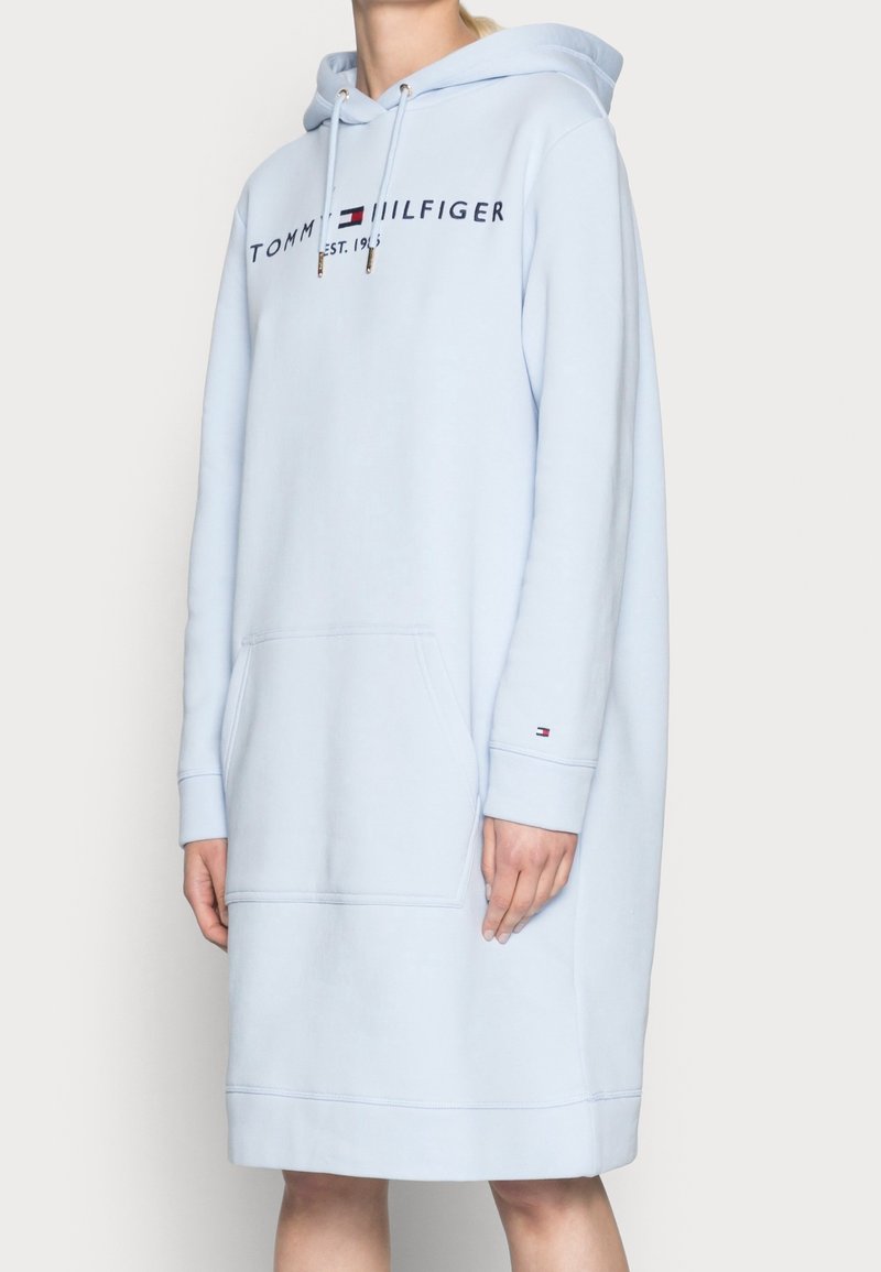 Lichtblauwe Tommy Hilfiger hoodie-jurk met een voorzak, logo-opdruk op de borst en klein logo op de manchetten van de linkerarm, gedragen door een persoon.