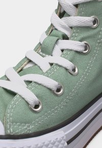 Gros plan d'une basket en toile vert menthe avec des lacets blancs passés à travers des œillets en argent et un bord de semelle en caoutchouc blanc visible.