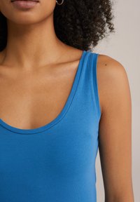 Srajca brez rokavov modre barve z ožjim izrezom, izdelana iz gladkega, elastičnega materiala. Ima oprijet silhueto in minimalne šive.