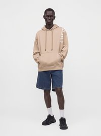 Homme portant un sweat à capuche beige avec un texte sur la manche, un short en jean, des chaussettes blanches et des baskets noires, debout devant un fond blanc uni.